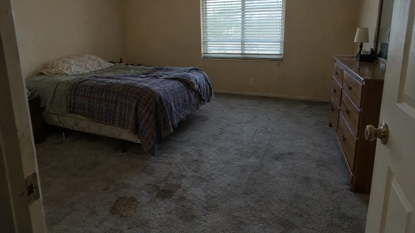 Stained Carpet Bedroom Before | Music City Floor Dimly lit bedroom with stained gray carpeting, a bed and a wooden dresser before new flooring